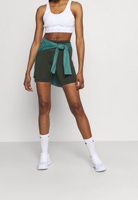 Vrouw in een witte sportbeha, donkergroene fietsbroek, een turquoise sweatshirt om haar taille gebonden, witte sokken en witte sneakers, staand op een grijze vloer.