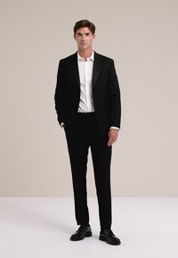 Fekete szmoking selyem hajtókával, fehér inggel párosítva. Slim-fit nadrág és fekete dresszcipő. Sima anyag, testre szabott dizájn.