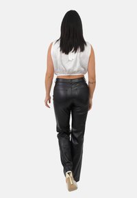 Donna con capelli scuri e lisci che si allontana, indossando un top bianco senza maniche, pantaloni in pelle nera e scarpe con tacco alto bianche.