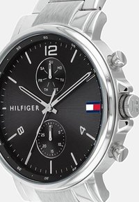 Reloj de acero inoxidable plateado con una esfera negra, que presenta subesferas de cronógrafo, marcadores de hora en negrita y un logotipo con acentos en rojo, blanco y azul.