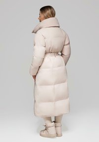 Žena v béžovém dlouhém péřovém kabátu se šňůrkou v pase a odpovídajících béžových botách, stojící zády k jednobarevnému šedému pozadí.