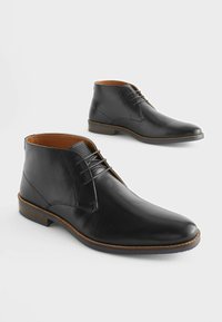 Botas de tobillo de cuero negro con un acabado suave, puntera redonda y diseño con cordones. Forro interior marrón y costuras en marrón contrastantes en la suela.