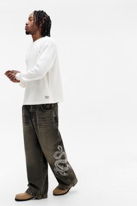 Fehér hosszú ujjú ing laza szabású, sötét olívazöld nadrággal, amelynek bal lábszárán skorpió grafika látható. A szettet bézs cipő egészíti ki.