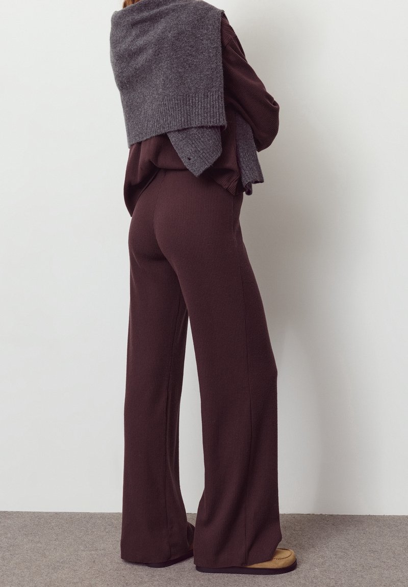 Personne portant un pantalon large côtelé marron foncé et un haut assorti, avec un pull gris drapé sur les épaules, debout contre un mur uni.