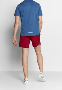 Persona vestida con una camiseta de mangas cortas azul, pantalones cortos deportivos rojos y zapatillas blancas, de pie contra un fondo blanco, vista desde atrás.