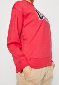 Czerwony sweter z okrągłym dekoltem, z wyraźnym granatowym haftowanym logo z przodu oraz ściągaczami przy mankietach i dole.