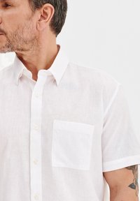 Biała lniana koszula z krótkim rękawem, z kołnierzykiem typu button-down i kieszonką na piersi. Materiał ma lekką, teksturowaną strukturę.