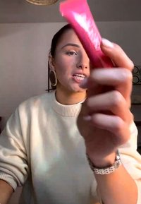 Jeune femme portant des boucles d'oreilles et un pull crème tient un tube de maquillage rouge et parle dans une pièce doucement éclairée.