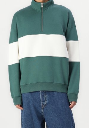 Person trägt einen grün-weiß farblich unterteilten Pullover mit halbem Reißverschluss, kombiniert mit locker sitzenden blauen Jeans vor einfachem Hintergrund.