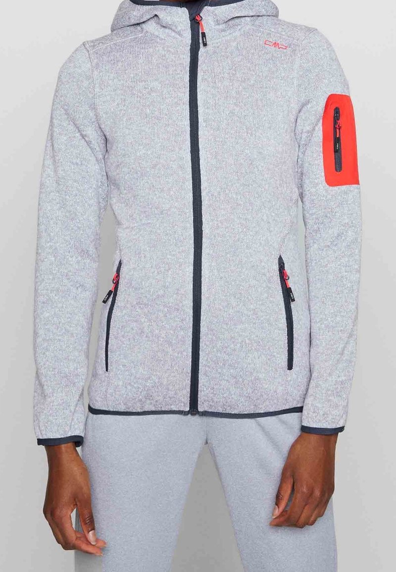 Grijze textuur zip-up hoodie met een capuchon, rode accentzak op de mouw en zwarte ritsdetails. Dressed met grijze broek.