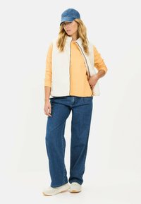 Hvid fleece vest over en gul langærmet skjorte, blå denim jeans og hvide sneakers. En blå kasket afslutter outfittet. Afslappet æstetik.