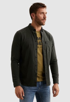 Homme portant une chemise vert foncé boutonnée par-dessus un t-shirt graphique couleur olive et un jean bleu, regardant vers la droite sur un fond uni.