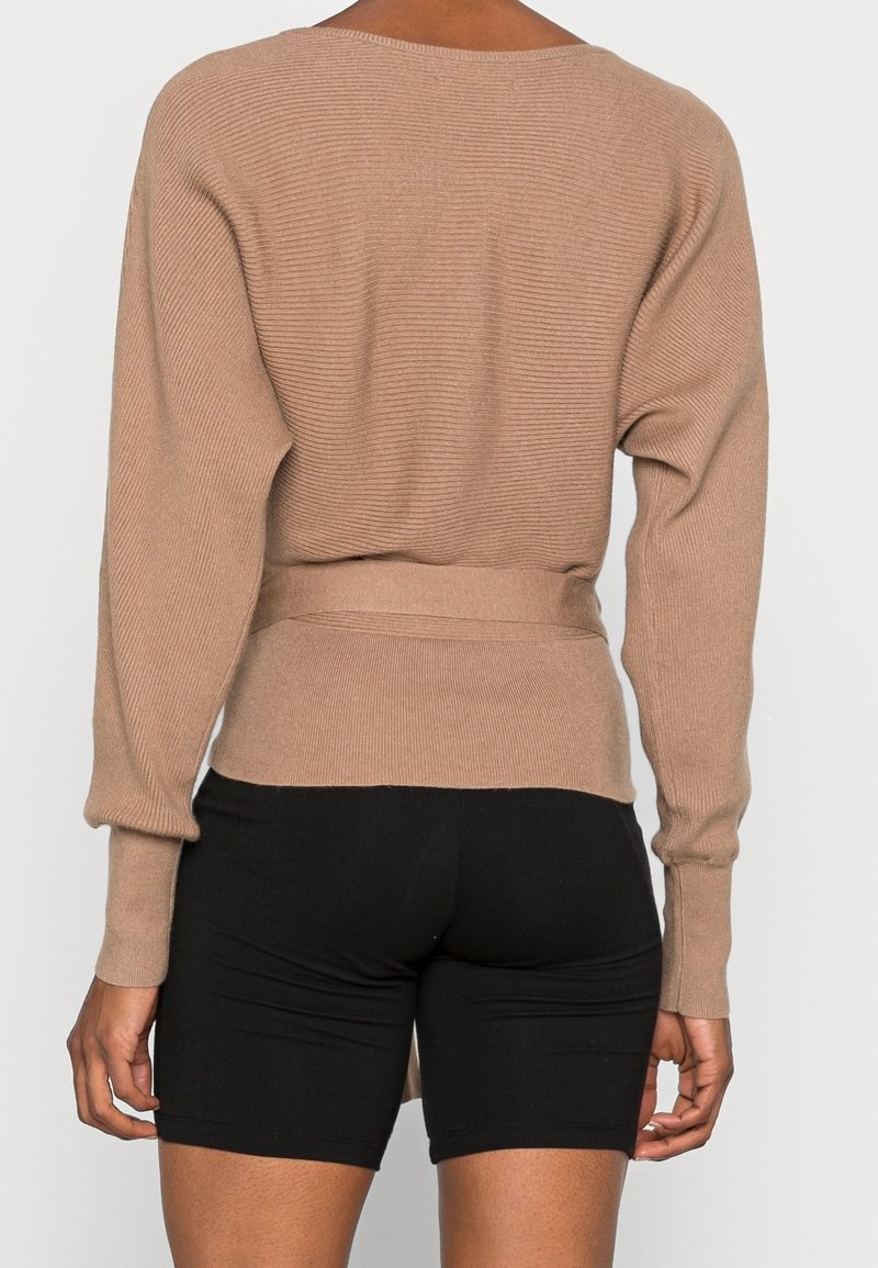 Personne portant un pull beige côtelé à manches longues avec une large ceinture et un short noir ajusté, vue de dos sur un fond blanc uni.