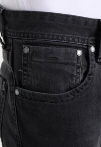 Jeans de mezclilla negra de talle alto, con herrajes plateados, un pequeño bolsillo frontal y detalles de costura sutiles.