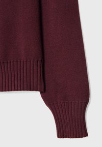 Manche longue et ourlet inférieur d'un pull en maille bordeaux foncé avec poignets et taille côtelés sur un fond clair.