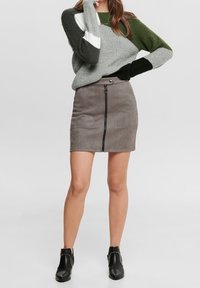 Mini-jupe en suède gris avec une fermeture éclair sur le devant, associée à un pull en maille vert et gris. L'ensemble comprend des bottines noires à rivets.