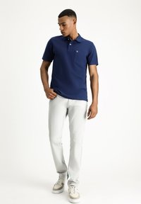 Polo shirt blu navy con tasca sul petto, abbinato a pantaloni grigi chiari e sneakers beige. Tessuto morbido, maniche corte e branding discreto.