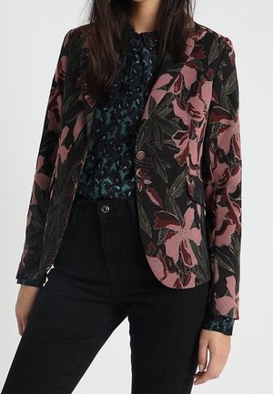 Vrouw draagt een zwart blazer met grote roze en groene bloemmotieven over een donkerblauw gebloemde blouse en zwarte broek.