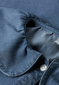 Jeansskjorta i mörkblått med en texturerad yta. Har en veckad krage och en vit knappdetalj.