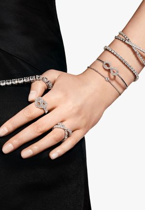 Zilverkleurige sieradenset op een hand omvat drie ringen met ingewikkelde ontwerpen, meerdere armbanden en sprankelende kristalaccenten.