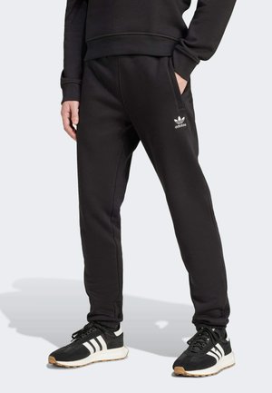 Persoon draagt zwarte Adidas joggingbroek en zwarte Adidas sneakers met witte strepen, staand met één hand in de zak.