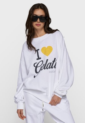 Kobieta w oversize białej bluzie z napisem "I [żółte serce] Gelati" i dopasowanych białych spodniach, pozująca w czarnych okularach przeciwsłonecznych.