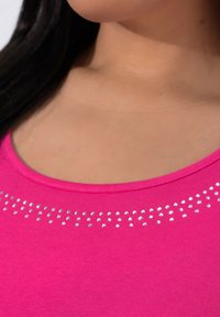 Lyserød skjorte i blødt stof med rund halsudskæring og en række små, skinnende rhinsten langs den øverste kant.