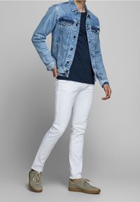 Muž v světle modré džínové bundě, námořnickém modrém tričku, bílých slim-fit kalhotách a béžových volnočasových teniskách stojící s jednou rukou v kapse.