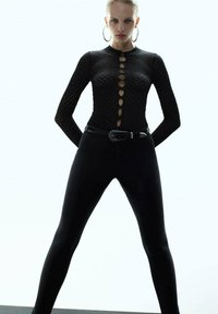 Top largo negro texturizado de manga larga con un cuello alto y detalle de botones en la parte delantera, combinado con leggings negros de cintura alta y un cinturón.