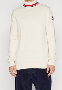 Cremefarbener Rollkragenpullover mit rot-navy gestreiftem Kragen, gerippten Bündchen und Saum, mit einem runden Logo-Patch am Oberarm.
