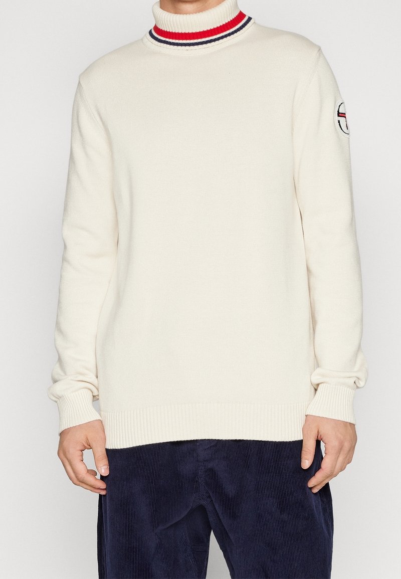 Cremefarbener Rollkragenpullover mit rot-navy gestreiftem Kragen, gerippten Bündchen und Saum, mit einem runden Logo-Patch am Oberarm.
