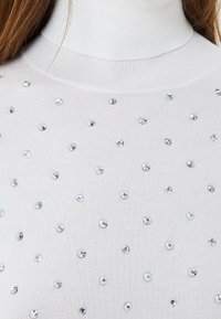 Weißer Strickpullover mit hohem Kragen, verziert mit gleichmäßig verteilten silbernen Pailletten für Textur und visuelles Interesse.