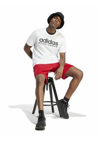 Camiseta de algodón blanca con el texto negro "adidas SPORTSWEAR", pantalones cortos rojos, sombrero de cubo negro, zapatillas negras, sentado en un taburete negro.