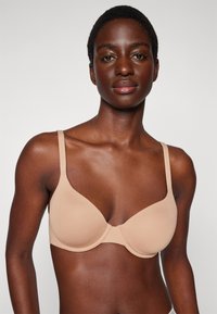 Beige gewatteerde beha met verstelbare bandjes, naadloos ontwerp en een gladde textuur, met een bescheiden plunge voorkant en geen zichtbare hardware.