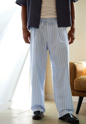 Man draagt losse lichtblauwe en witte verticale gestreepte broek met trekkoord, zwarte schoenen, donker shirt en beige top binnenshuis.