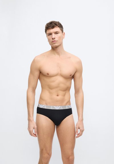 Άνδρας χωρίς μπλούζα, με κοντά σγουρά μαλλιά, φοράει μαύρα εσώρουχα με γκρι ελαστική ζώνη μέσης που έχει λευκό λογότυπο, στέκεται μπροστά σε λευκό φόντο.