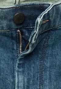 Primer plano de unos pantalones vaqueros de mezclilla azul con una cremallera metálica parcialmente desabrochada y un botón negro etiquetado "MNG DENIM".