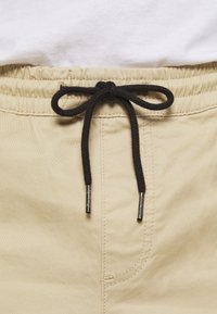 Beige shorts with elastic waistband, featuring a dark brown drawstring and metal aglets, and a side seam pocket detail.