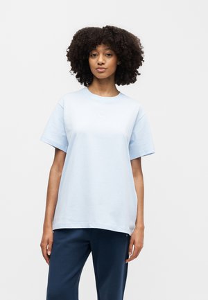 Femeie cu păr creț purtând un tricou Nike albastru deschis și pantaloni bleumarin, stând în picioare pe un fundal alb simplu.