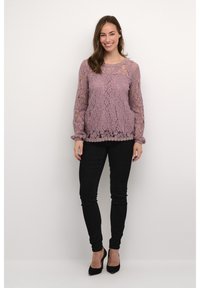 Spitzenoberteil mit langen Ärmeln in hellem Lila, mit einer gewellten Ausschnittlinie und Rüschensäumen, kombiniert mit schwarzen Skinny-Jeans und High Heels.