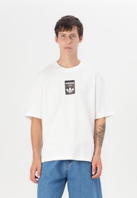 White cotton t-shirt with short sleeves, featuring a black and leopard print Adidas logo patch centered on the chest. Casual fit.