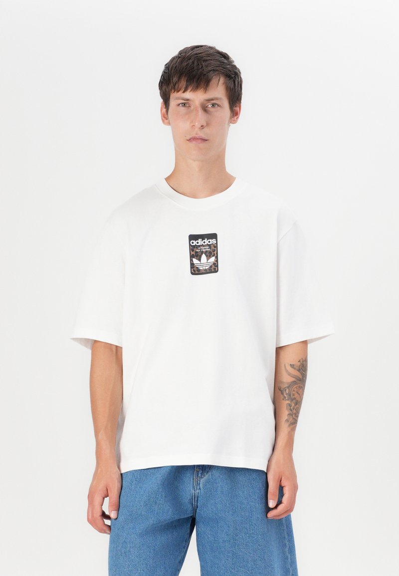 White cotton t-shirt with short sleeves, featuring a black and leopard print Adidas logo patch centered on the chest. Casual fit.