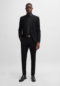 Traje negro con diseño ajustado, que presenta solapas de muesca, mangas largas, camisa de vestir a juego y corbata. Cinturón de piel y zapatos pulidos.