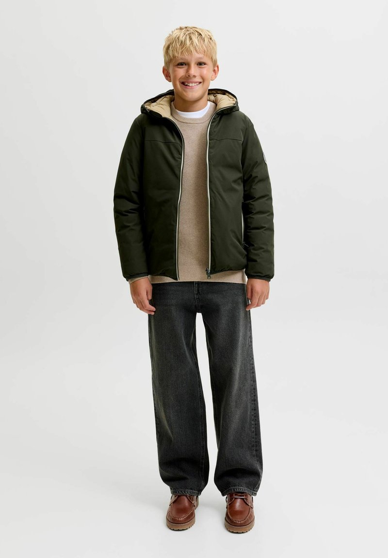 Garçon blond souriant portant une veste à capuche vert foncé, un pull beige, un jean noir et des chaussures marron, debout sur un fond blanc uni.