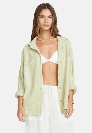 Vrouw draagt een lichtgroen oversized overhemd over een witte bikinitop en bijpassende witte broek met trekkoord, afgewerkt met een kralenketting.