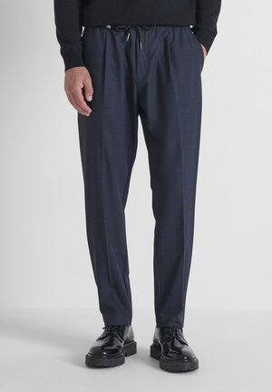 Man in marineblauwe, geruite, op maat gemaakte broek met een drawstring taille en zwarte, gepolijste veterschoenen, staande voor een effen achtergrond.