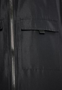Jachetă neagră cu fermoar, dotată cu două buzunare mari pe piept, având un material texturat și un model vertical cu dungi. Accente din metal argintiu pe fermoar.