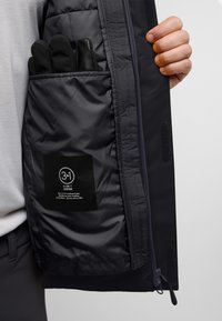 Intérieur d'une veste 3-en-1 gris foncé avec une poche zippée contenant des gants noirs. Comprend une étiquette d'entretien avec des informations sur les caractéristiques de protection contre les intempéries.