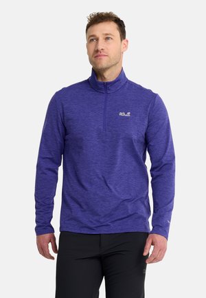 Man met een paarse lange mouwen zip-up top en zwarte broek, staat en kijkt iets omhoog met een neutrale gezichtsuitdrukking.