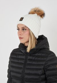 Donna che indossa un berretto bianco in maglia con un pompon marrone e una giacca a vento nera con colletto alto, rivolta a sinistra su uno sfondo bianco.